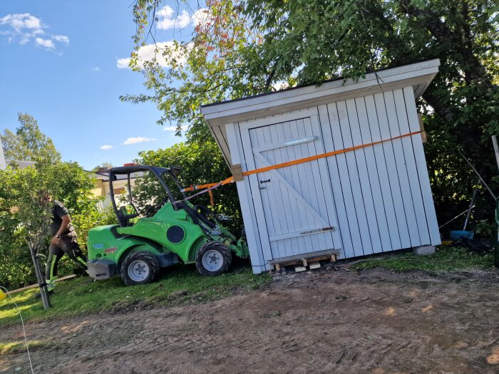 Grön minilastare bredvid lutande vedbod på en gräsmatta, omgiven av träd. En person står i bakgrunden.