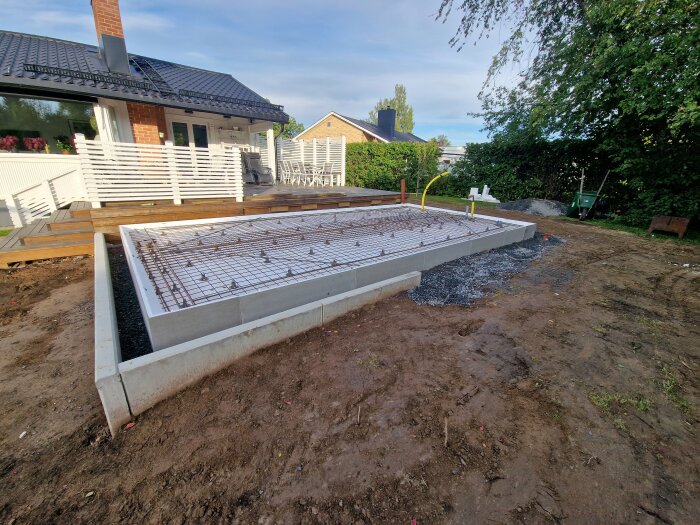 Grundplatta för attefallshus under konstruktion på lutande tomt, med L-stöd och armeringsjärn intill hus med altan.