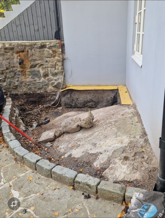 Sluttande jord och stenar bredvid en husgrund med gula isodränplattor synliga, omgiven av en murad kant och stenläggning.