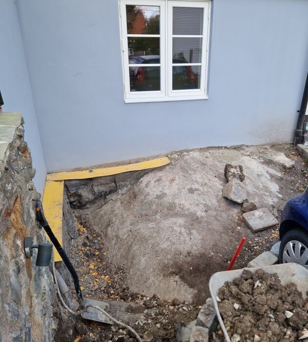 Utgrävd rabatt intill husgrund med berggrund synlig. Isodränplattor längs väggen, gula brädor på marken, stenblock och verktyg runt området.