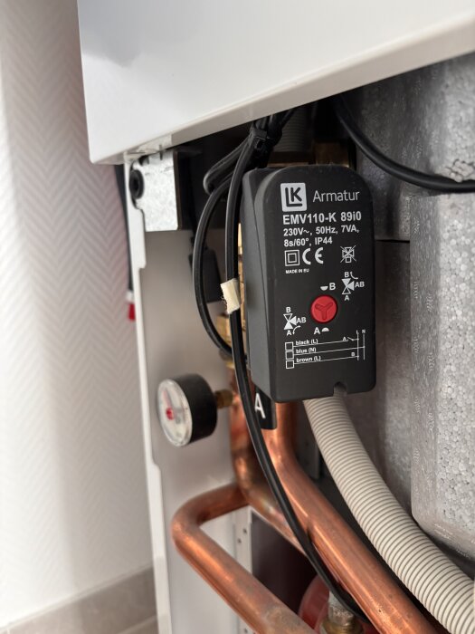 Värmesystemkomponent med LK Armatur EMV110-K 8910 och kopparrör i närbild, visar elanslutningar och en tryckmätare.