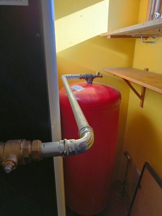 Röd ackumulatortank i ett gult rum, ansluten till en metallrörledning. På väggen syns hyllor och ett fönster.