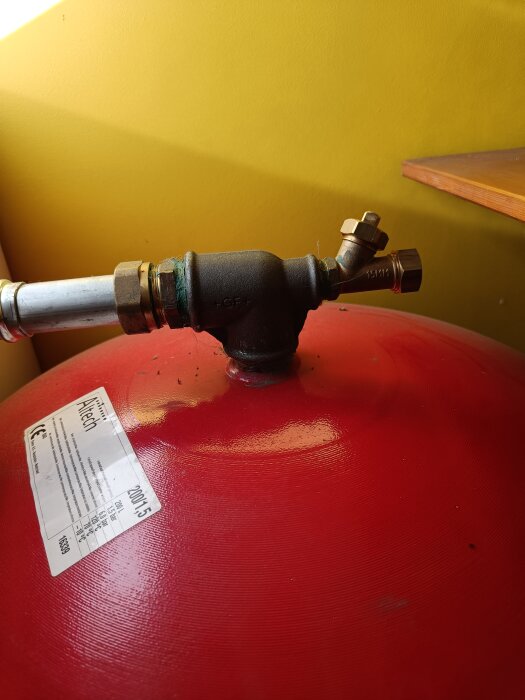 Röd ackumulatortank med ett kopparrör och ventil i en villa; mot en gul vägg och träyta i bakgrunden.