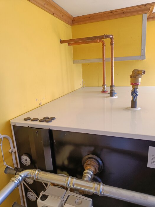 Ackumulatortank med installation av rör och ventiler synliga ovanpå en vit yta mot en gul vägg i ett hus.