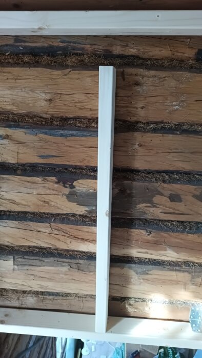 Trävägg med horisontella stockar och en vertikal bräda i ljusare träfärg, isolering mellan stockarna.