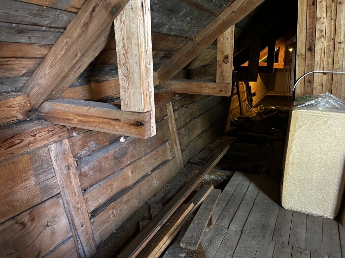 Vind med exponerade takstolar och liggande timmer i ett gammalt hus, med lutande yttervägg. Perspektiv ger illusion av större lutning.