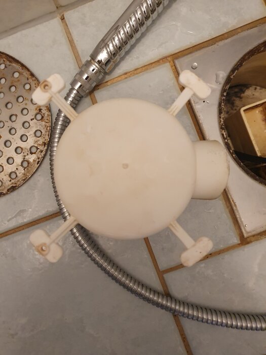 Plastkåpa från duschbrunn med fyra plastflikar, intill rostig brunn och duschslang på kakelgolv i äldre badrum.