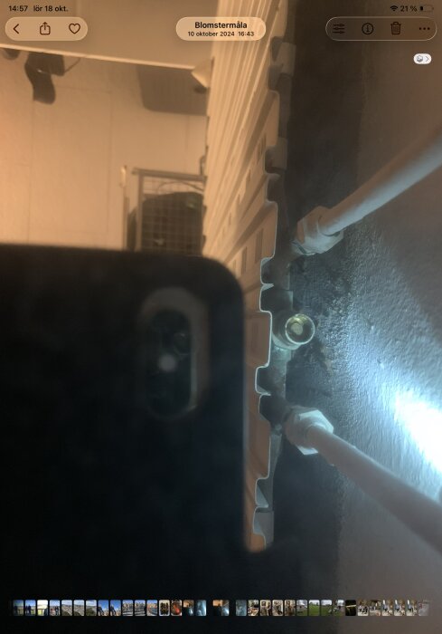 Närbild av en del av en radiator med rör och en koppling i ett svagt upplyst rum. Reflektion från en telefon syns i förgrunden.