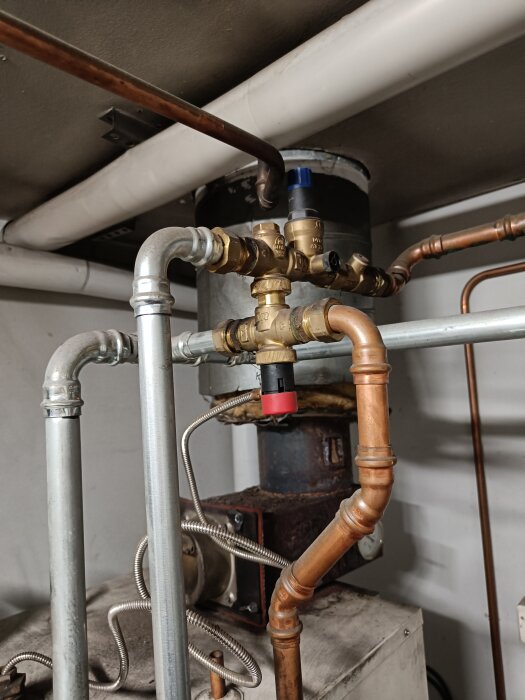 Övertrycksventil på vedpanna med kopparrör, röd hatt, blå ratt och anslutna rör, del av ett värmesystem i pannrum.