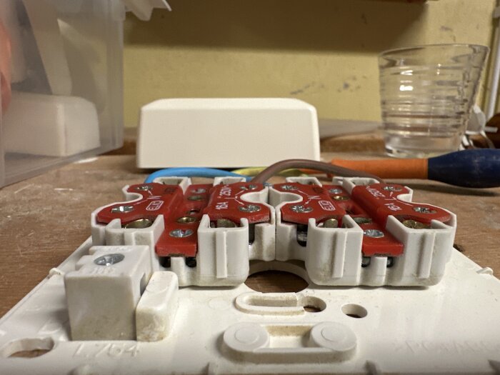 Elektrisk kopplingsplint med röda skruvterminaler på arbetsbänk; plats för flera trådar syns, inramad av vit plast.