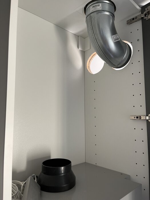 Ventilationsrör i metall med böjning, monterat i taket på ett vitt skåp. Svart adapter och elkablar synliga på hyllan nedanför.
