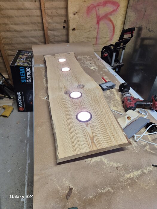 Ett trästycke med fyra infällda lampor ligger på ett arbetsbord, omgivna av verktyg och sågspån i en verkstad.