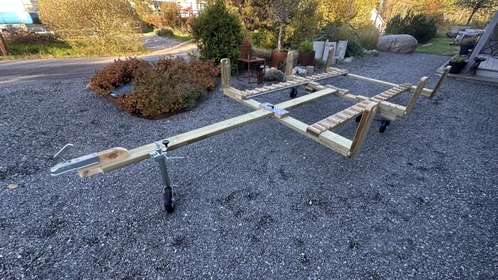 Hemmagjord båtvagn på grusplan med trä- och metallelement, omgiven av buskar och stenar i en trädgårdsmiljö.