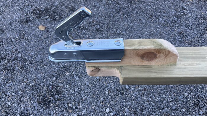 Bild på en träbalk med en monterad dragkrok av metall för båtvagn, liggande på grus.