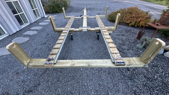 Trävägn med metallarmar och hjul, byggd för trampbåt, stående på grus framför ett hus och omgiven av buskar och trädgårdsdekorationer.