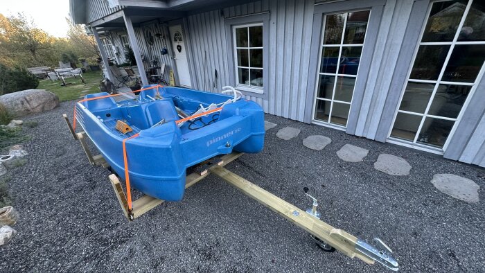 Blå trampbåt på en ny båtvagn framför ett hus. Båtvagnen har justerbar dragstång och är måttanpassad för båten.