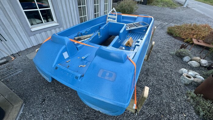 Blå trampbåt placerad på en hembyggd båtvagn, omgärdad av orange remmar, stående på en grusplan bredvid ett hus.