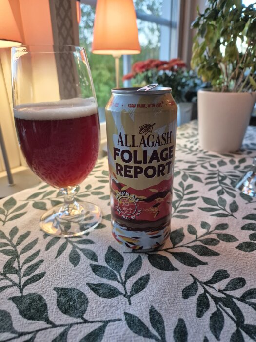 Burk och glas med rödbrun Allagash Foliage Report Autumn Ale på ett dukat bord med bladmönster, omgiven av växter och belysning.