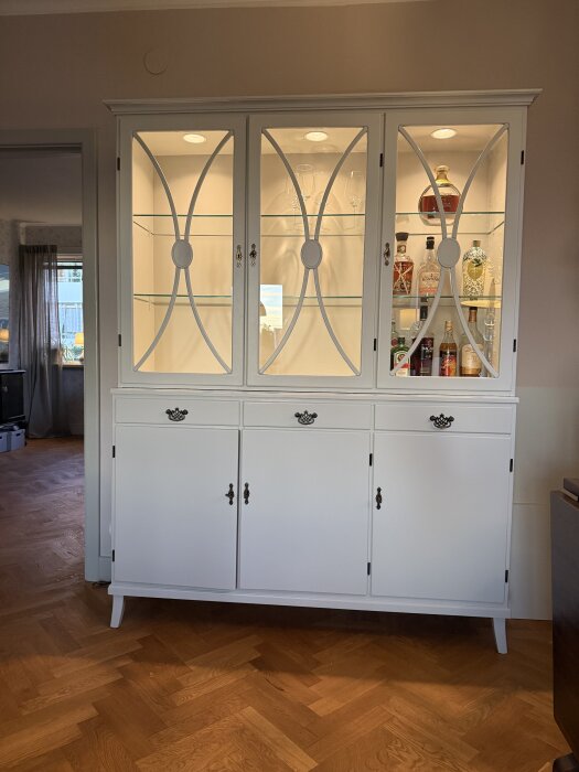 Vitt vitrinskåp med glasdörrar och belysning, innehållande spritflaskor och glas.