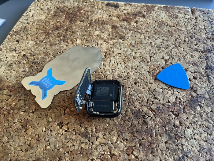 En öppen smartklocka på en korkmatta, omgiven av verktyg för batteribyte.