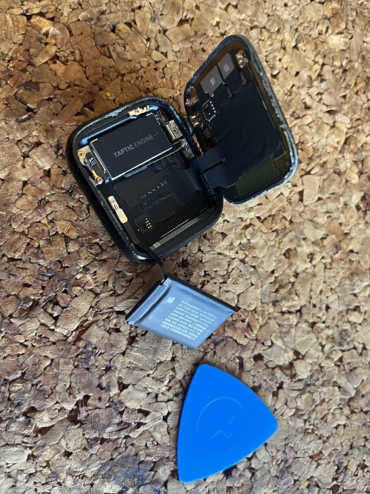 En öppen smartklocka med synlig batteribyte, liggande på en korkmatta med verktyg för öppning.