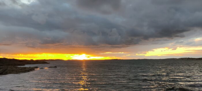Solnedgång över havet med dramatiska moln och reflektioner av det gyllene ljuset på vattnet. Klippor syns vid horisonten.