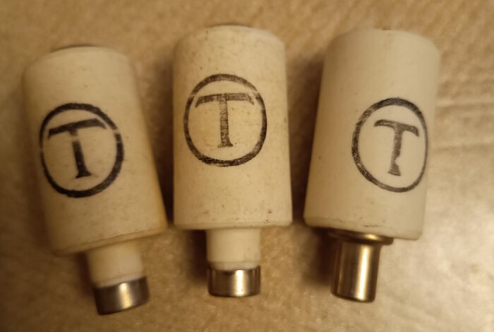 Tre äldre tröga säkringar med bokstaven T i en ring, ett äldre symbol för tröghet, placerade på ett bord.