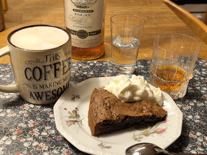 En kladdkaka med grädde på tallrik, cappuccino i mugg bredvid Bowmore whisky och glas med vatten, stående på blommig duk.