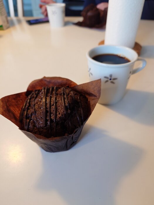 Chokladmuffin med chokladglasyr på ett bord bredvid en kopp kaffe.