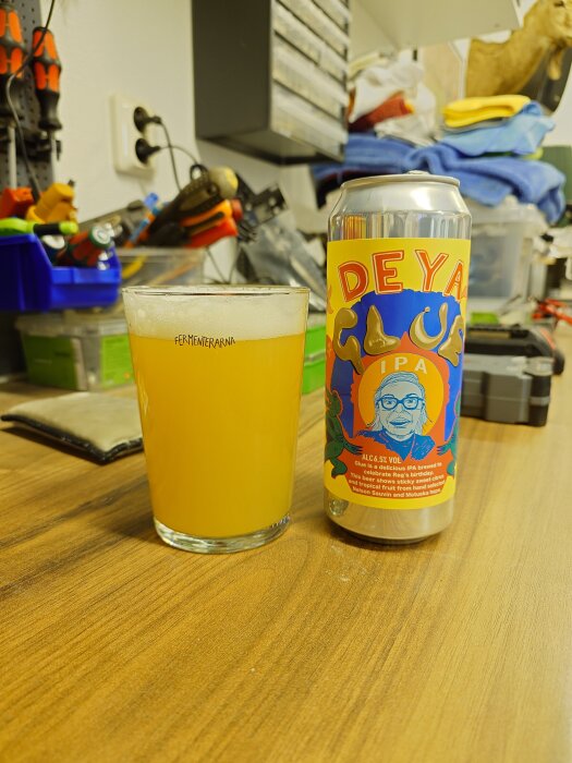 Deya Glue IPA-burk och glas med öl på en arbetsbänk med verktyg i bakgrunden.
