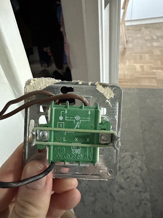 Grön elektronisk strömbrytare med brun och svart kabel kopplad, monterad på en metallplatta, håller på att installeras i ett hem.