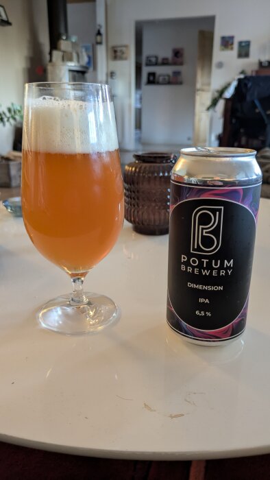 Ölglas fylld med orange IPA och en ölburk från Potum Brewery på ett vitt bord i ett vardagsrum.