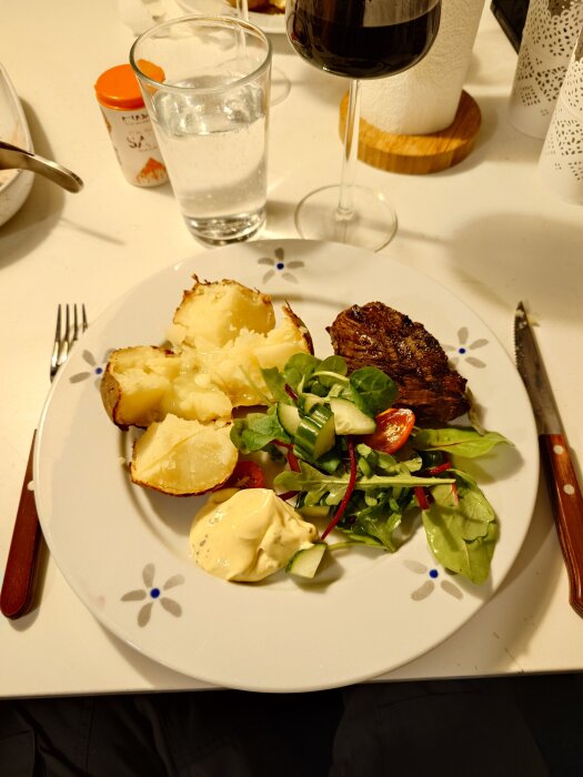 Bakad potatis, grillad rostas, sallad och bearnaisesås på en tallrik med ett glas rödvin och vatten bredvid.
