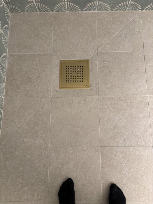 Beige klinkergolv med ny fog och guldfärgad golvbrunn, flätmönstrade väggar syns delvis, sockor i bildens nederkant.