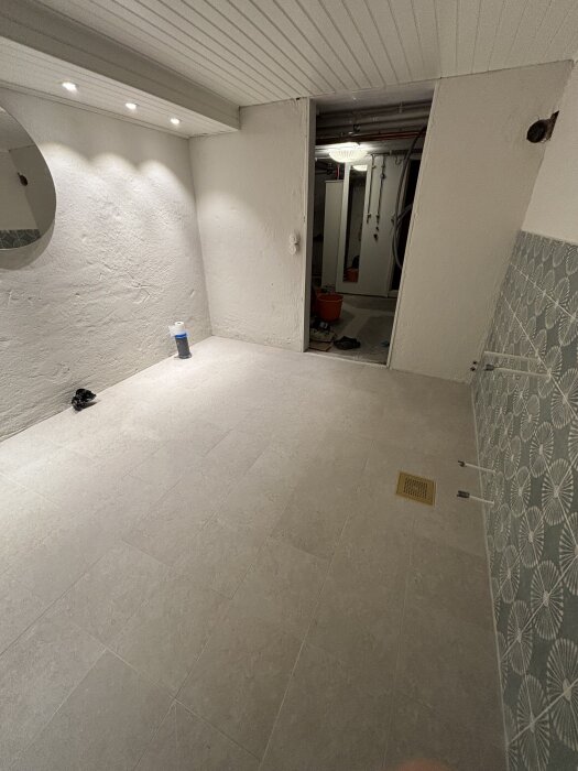 Nyfogad badrumsgolv med kakel, en spegel på väggen och ett materialrör i hörnet, redo för installation av vitvaror och duschvägg.