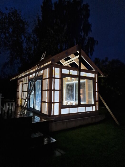 Byggprojektets stomme med limträ, stora fönster och skjutdörr visas i nattligt ljus med stege lutad mot väggen.