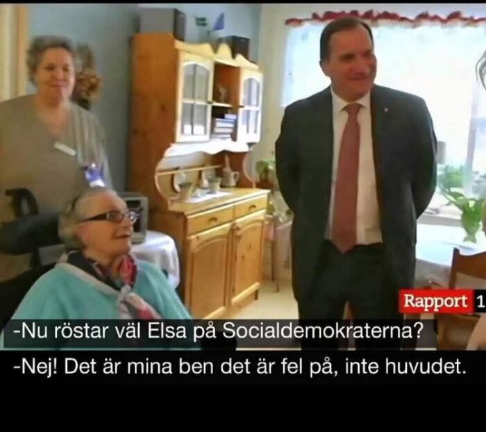 En äldre kvinna i rullstol samtalar med en man i kostym i ett rum med träinredning. Kvinna i bakgrunden står bredvid en byrå. Text från tv-sändning syns.