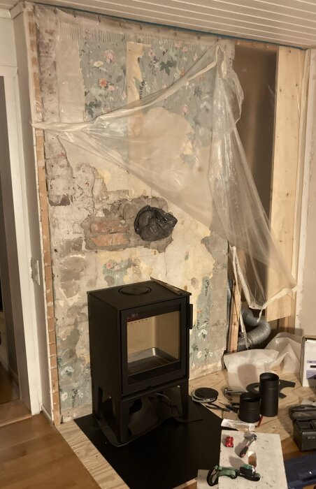 Murad vägg med synlig rökkanal, delvis täckt av plast, ny svart kamin på golvet, byggmaterial och verktyg runt.