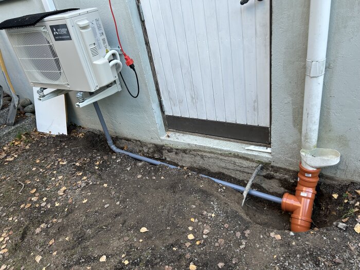 Luftvärmepump på vägg bredvid dörr, med grått rör anslutet till orange rör för kondensavledning, omgiven av grus och löv.