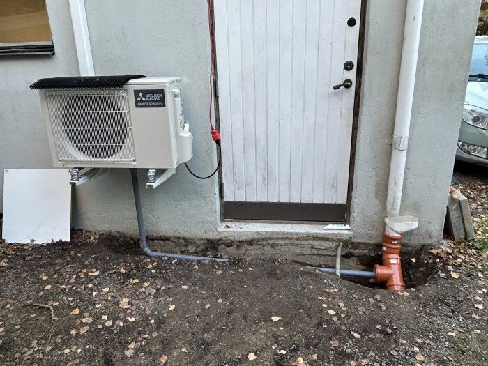 Värmepump installerad utomhus på husvägg med grå rör för kondensavledning nedgrävt i marken, intill en dörr och orange dräneringsrör.