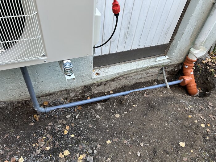 Grå ventilationsrör från en luft/vatten-värmepump installerat i marken bredvid en orange avloppsledning och en vit byggnadsvägg.