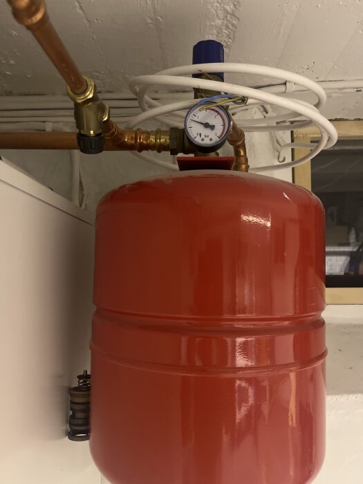 Röd expanstionskärl med tryckmätare och kopparrör i en källare.