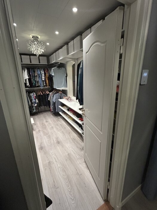 Klädkammare med klädhängare och hyllor fyllda med kläder, skoboxar ovanpå och en ljuskrona i taket.