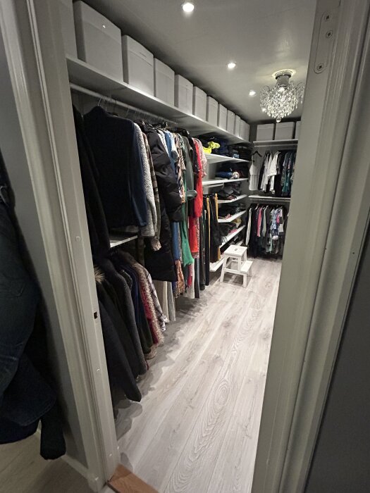 Klädkammare med kläder hängande på stång, lådor ovanpå hyllor och kristallkrona i taket, nyligen renoverad och organiserad.