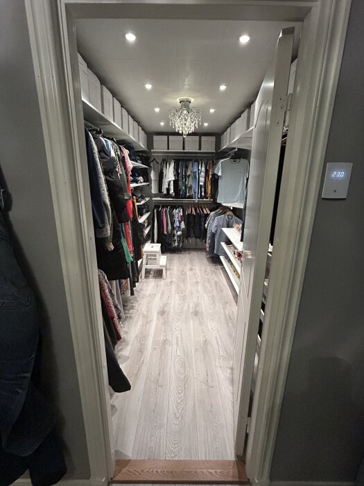 Modern walk-in closet med avslutade klädhyllor, kläder organiserade på galgar, ljus parkettgolv och kristallkrona i taket.