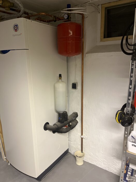 Värmepanna med expansionskärl monterat ovanpå. Rörledningar och tryckmätare synliga. Hörnet av ett rum med hylla och hörlurar.