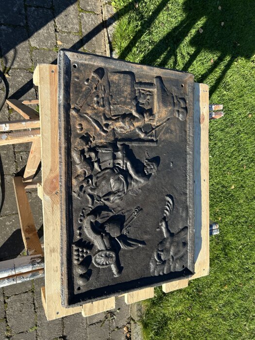 Gammal dekorativ sättungsplatta i svart metall med ingraverade mönster, placerad på träyta utomhus för restaurering.