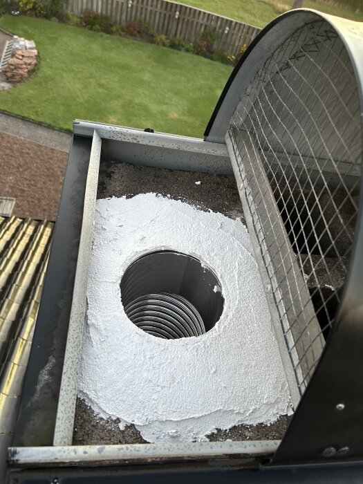 Kaminens murstock tätad med vit fogmassa, ventilerad via en metallinsats, med trädgård i bakgrunden.