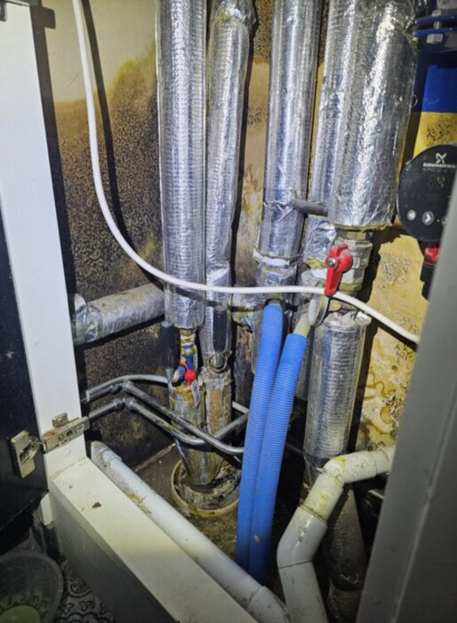 Fjärrvärmesystem med isolerade rör och ventiler i ett litet schakt, med synlig vägg och golv i bakgrunden, tvättstugeprojekt.