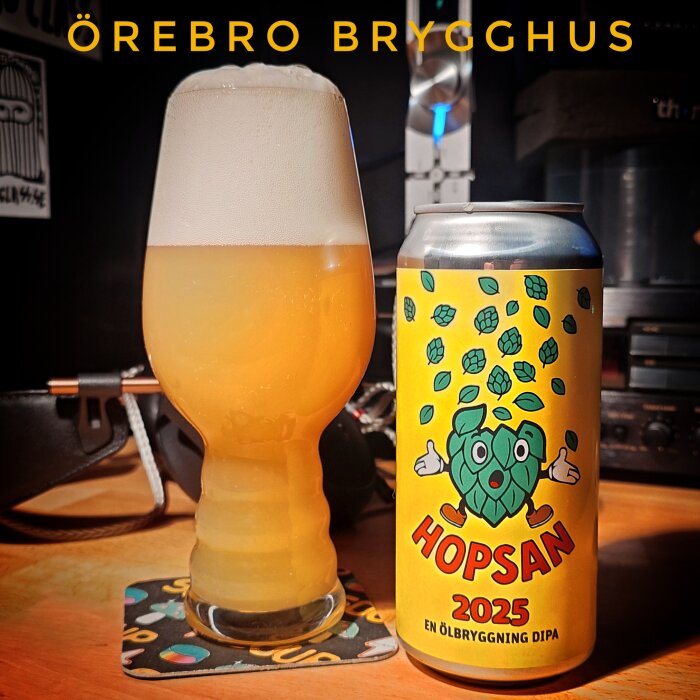 Ölglas och burk av Hopsan 2025 DIPA från Örebro Brygghus på bord med musikrelaterad bakgrund.
